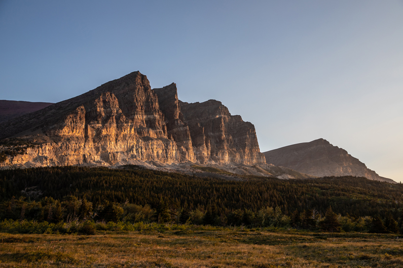 Low-angle sunlight illuminates a cliff-face, perched above sloping coniferous forest and a grassland below.