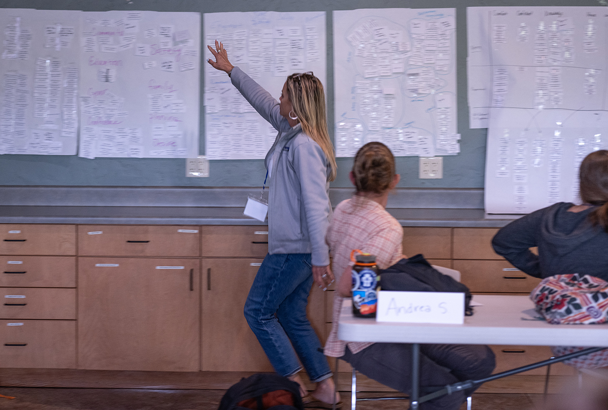 Woman gestures at posters containing lists of information while she is walking across a room full of conference participants..