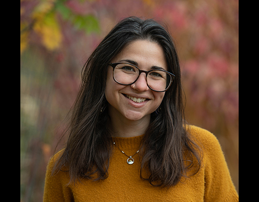 Woman with dark hair, glasses and wearing a yellow sweater smiles at camera.
