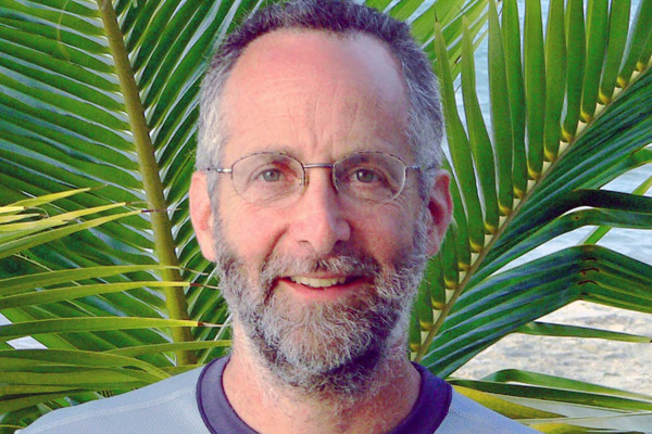 Bearded man, wearing glasses, smiles at the camera while standing before a spray of palm leaves.